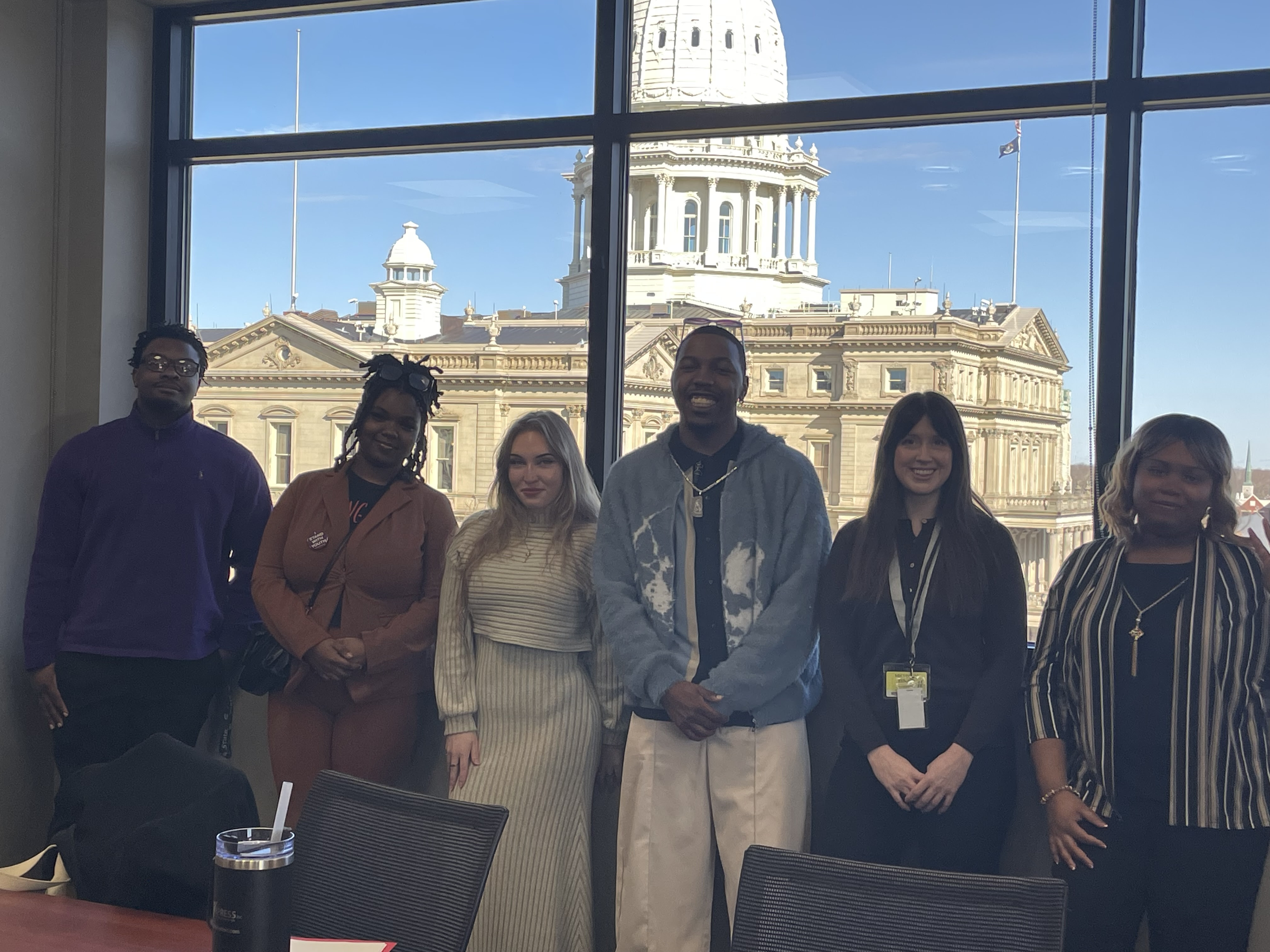 Six youth advocates standing together with the Michigan State Capitol dome visible through the window behind them