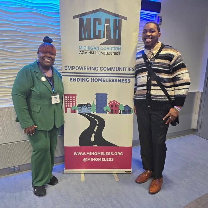 Two advocates smiling next to a Michigan Coalition Against Homelessness banner reading 'Empowering Communities, Ending Homelessness'