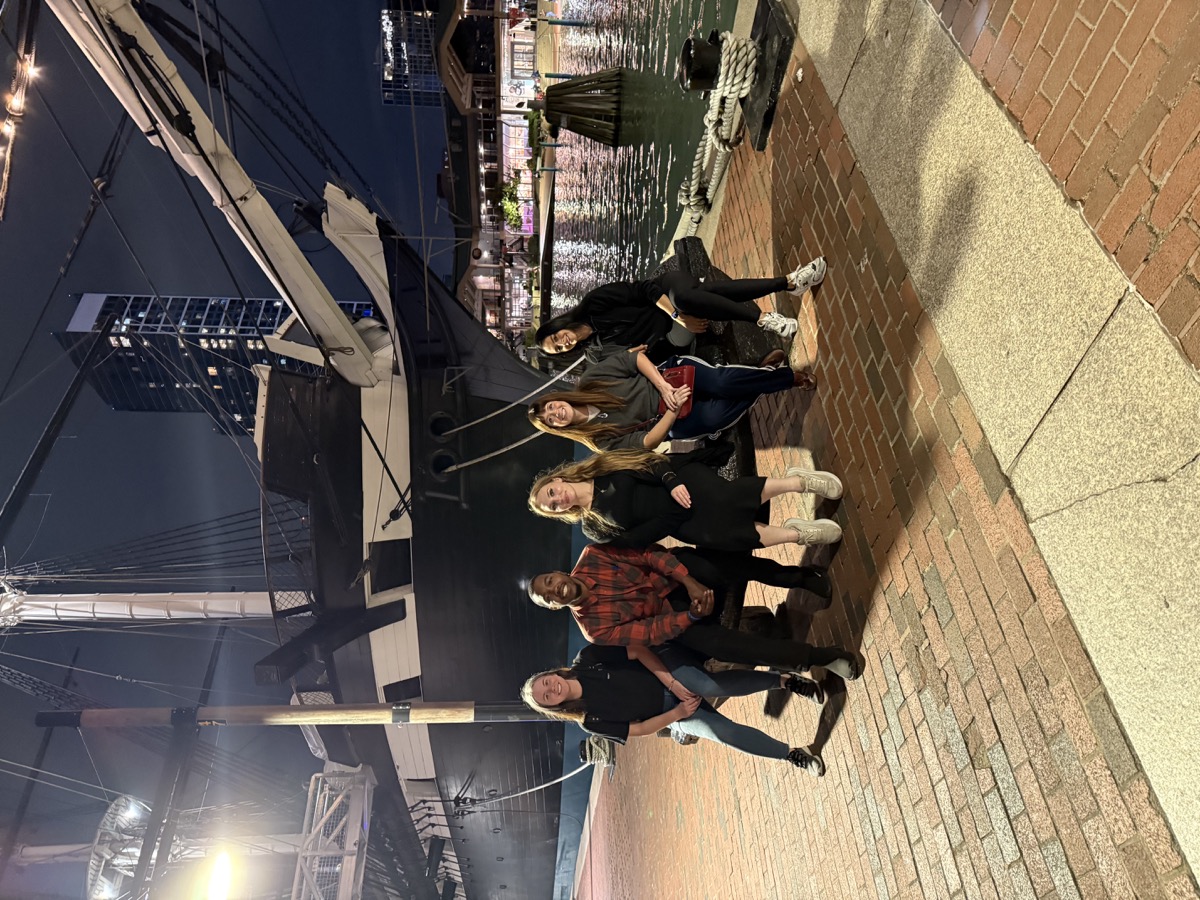 Five team members smile together at the Baltimore waterfront near a historic ship during an evening conference outing