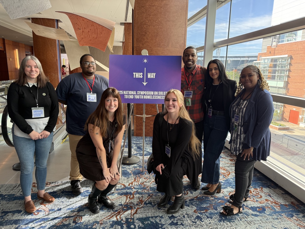 Team members at the National Symposium to End Youth Homelessness, standing in front of the event's purple signage